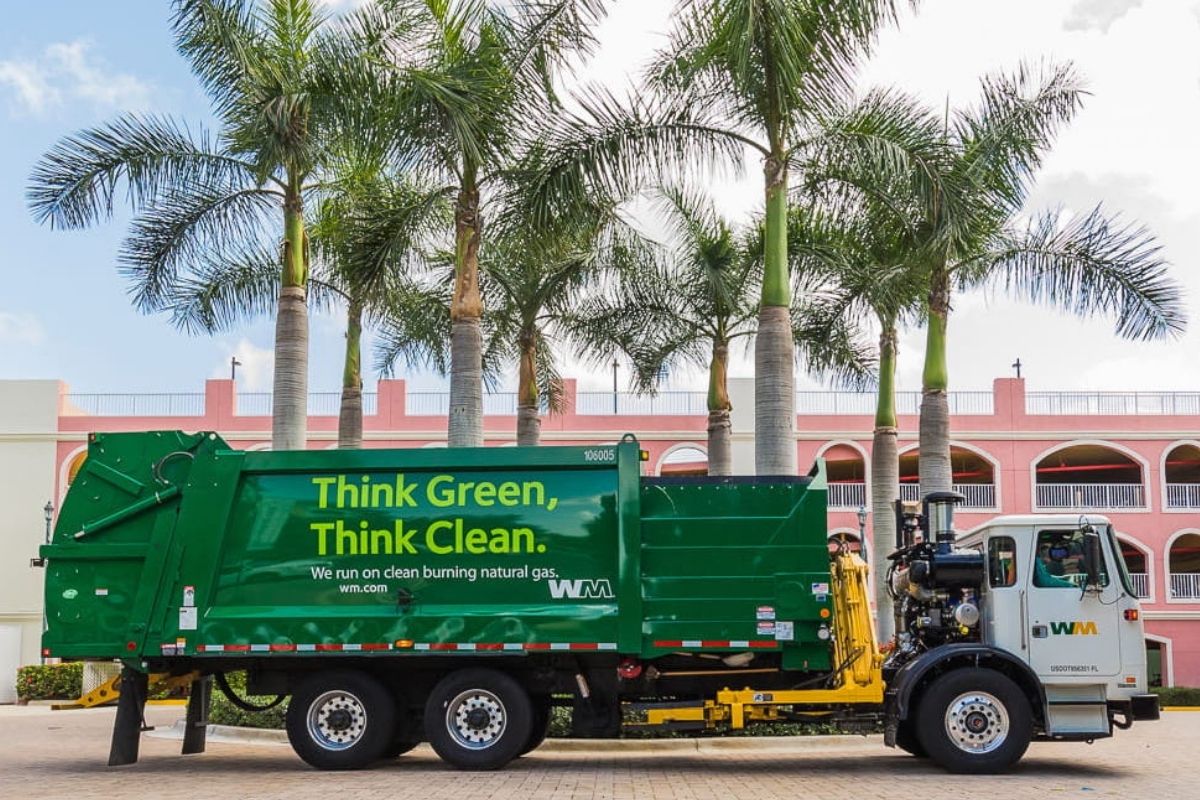 A WM garbage pickup truck parked in front of some palm trees in Mango, Florida with a pink building in the background.