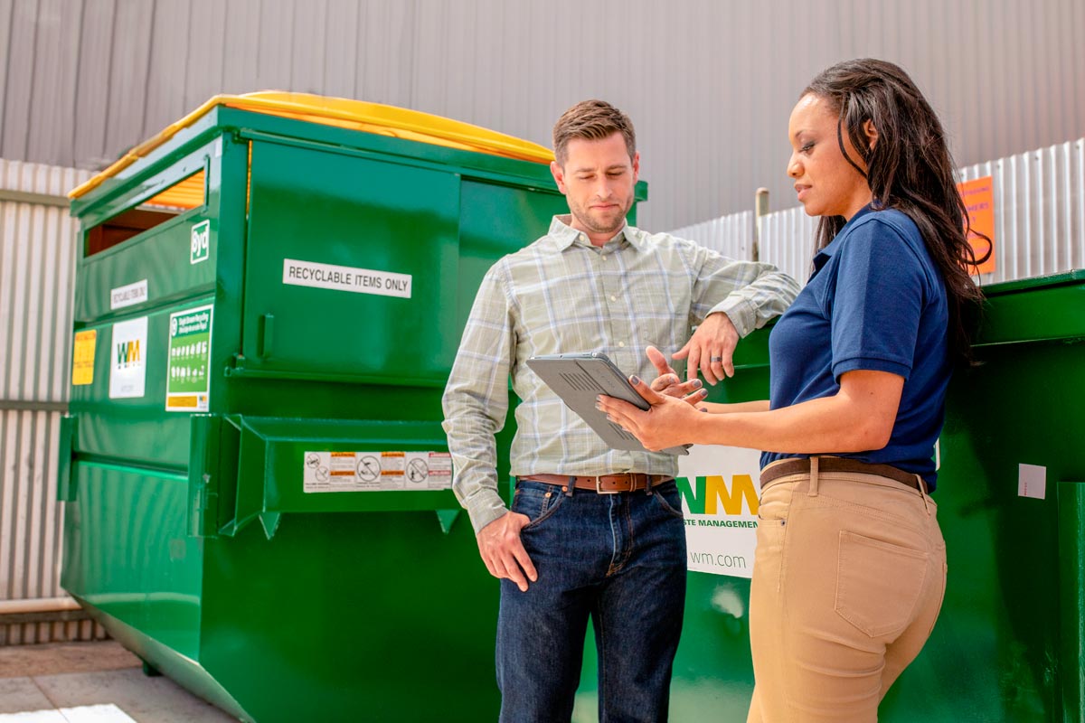 A man and a woman look at a tablet while standing outside near a Waste Management dumpster and recycling container.